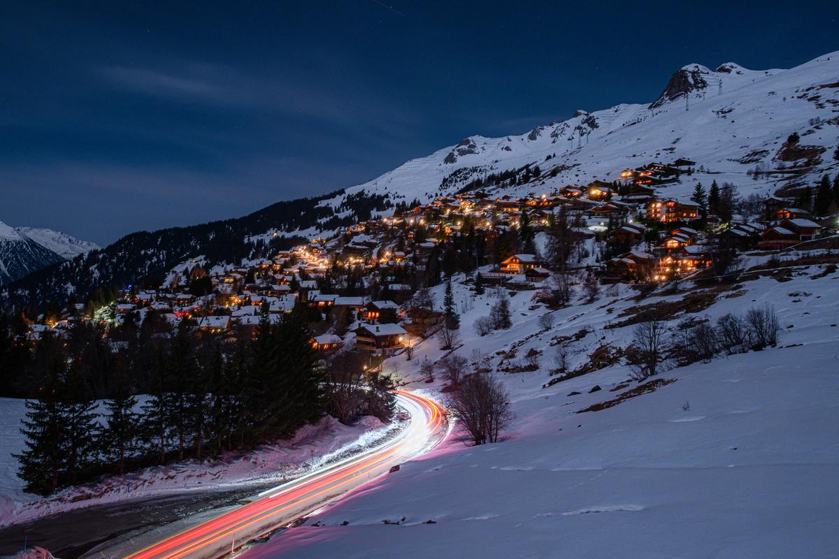 Long exposure of car lights tracing a red and white curve along a snowy mountain road, with lit chalets on the slope above at night