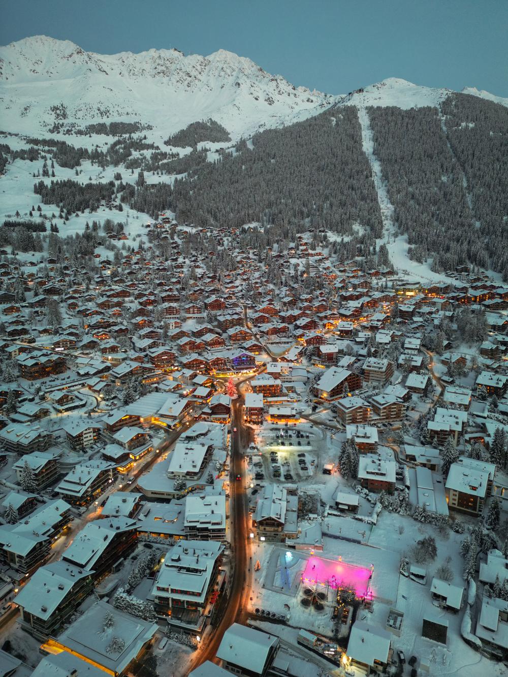Aerial view of Verbier at dusk after snowfall, the main road lit orange between snow covered chalets, with the mountains and a forest piste behind