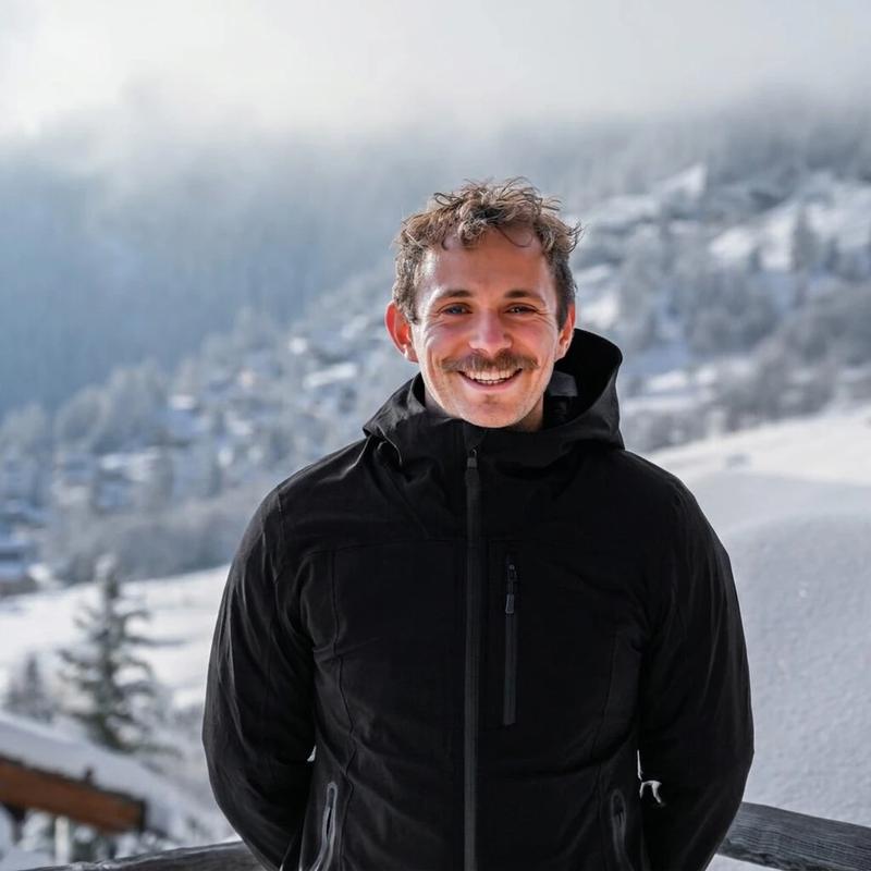 Will Stirrup, founder of Alpine Chauffeur Club, smiling in a black jacket on a snowy balcony with chalets and pine trees behind